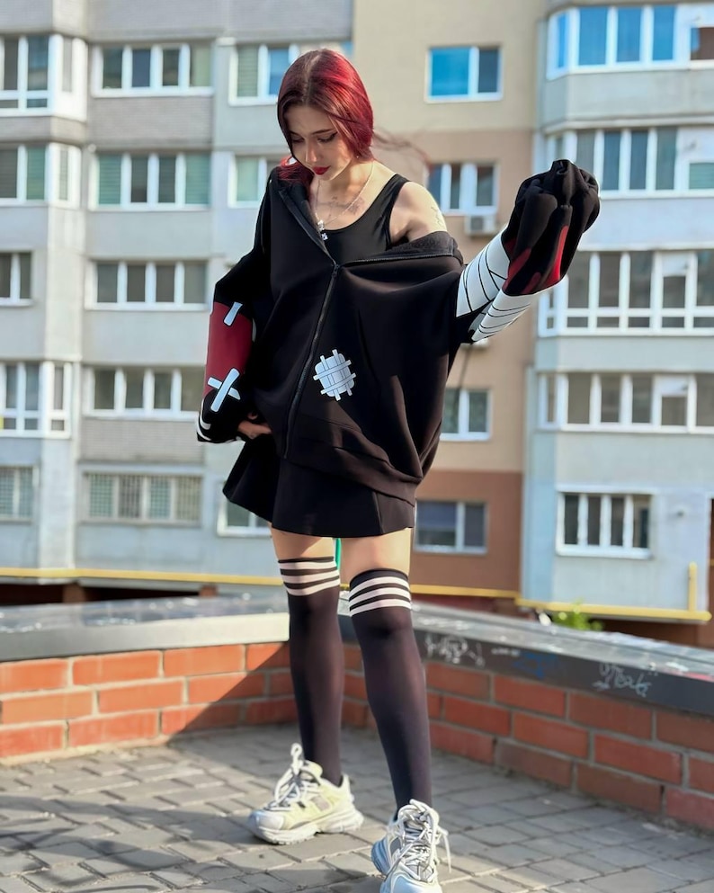 Op de afbeelding: Zwarte hoodie met rits met rode en witte accenten op de mouwen. De hoodie heeft een witte patch met een tandwielontwerp. De outfit omvat een zwarte rok, zwarte kniehoge sokken met witte strepen en witte sneakers.