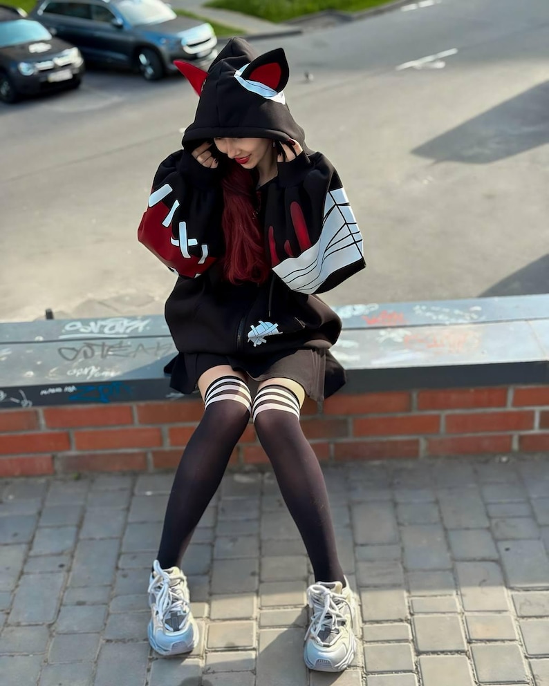 Op de afbeelding: Zwarte hoodie met kattenoren en rode accenten. De mouwen hebben witte en rode details. De persoon draagt zwarte dijhoge sokken met witte strepen en grijze sneakers.