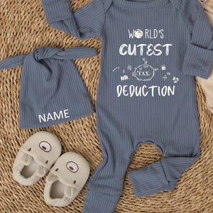 May include: A blue ribbed baby outfit set with a matching hat and slippers. The outfit includes a long-sleeved onesie with the text "WORLD'S CUTEST TAX DEDUCTION". The hat has the word "NAME" printed on it. The slippers are beige.
