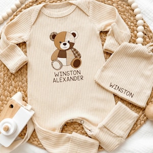 May include: A cream-colored ribbed long-sleeve baby romper and matching knotted hat. The romper features a brown patchwork teddy bear graphic with the text WINSTON ALEXANDER printed below. The hat features the name WINSTON printed in brown.