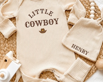 May include: A beige ribbed long-sleeve romper and matching knotted hat set. The romper features the brown text "LITTLE COWBOY" above a small brown cowboy hat icon. The hat is personalized with the brown text "HENRY" printed on the front.
