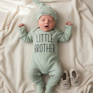Pode incluir: Um rec&eacute;m-nascido vestindo um macac&atilde;o de malha canelada verde s&aacute;lvia com "LITTLE BROTHER" impresso em preto. O beb&ecirc; tamb&eacute;m usa um chap&eacute;u de n&oacute; combinando com o nome "Thomas" e botinhas bege em forma de urso. O beb&ecirc; est&aacute; deitado em um cobertor creme.