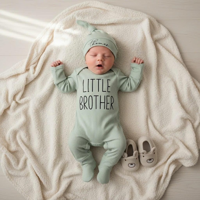 Pode incluir: Um rec&eacute;m-nascido vestindo um macac&atilde;o de malha canelada verde s&aacute;lvia com "LITTLE BROTHER" impresso em preto. O beb&ecirc; tamb&eacute;m usa um chap&eacute;u de n&oacute; combinando com o nome "Thomas" e botinhas bege em forma de urso. O beb&ecirc; est&aacute; deitado em um cobertor creme.