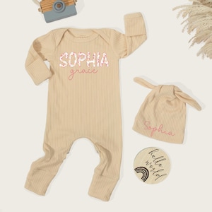 Pu&ograve; includere: Una tutina a maniche lunghe a coste color beige con il nome Sophia Grace stampato in caratteri floreali e corsivi. Include un berretto annodato abbinato con il nome Sophia e un disco di legno rotondo con il testo hello world.