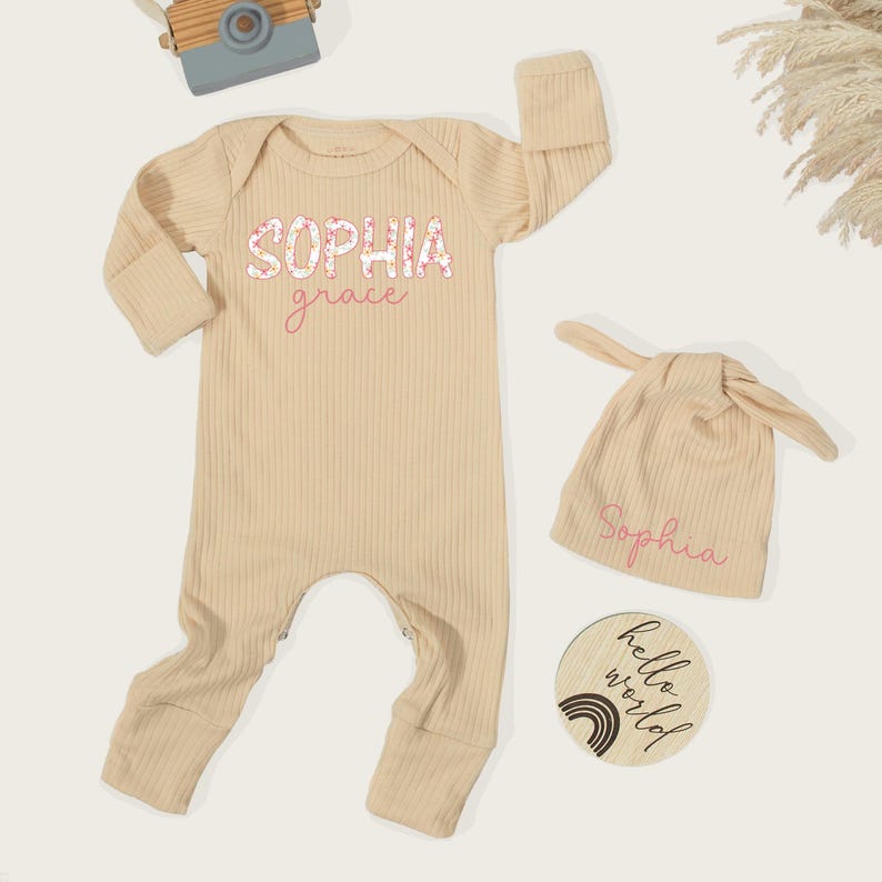 Pu&ograve; includere: Una tutina a maniche lunghe a coste color beige con il nome Sophia Grace stampato in caratteri floreali e corsivi. Include un berretto annodato abbinato con il nome Sophia e un disco di legno rotondo con il testo hello world.