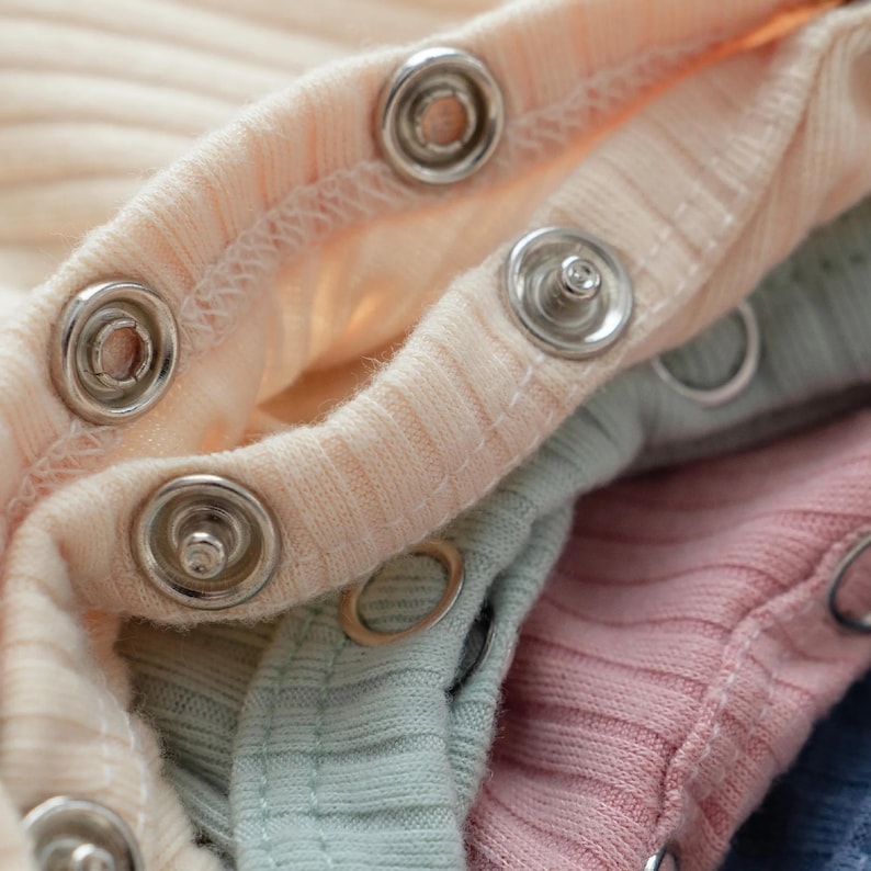 Pu&ograve; includere: Una pila di capi in tessuto a coste con chiusure a pressione metalliche. Lo strato superiore &egrave; di colore crema chiaro, seguito da strati in verde salvia tenue, rosa polvere e un tocco di blu scuro, con cuciture visibili attorno ai bottoni.