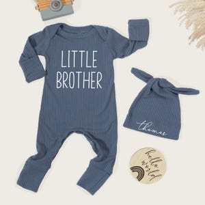 Pode incluir: Um conjunto de roupa de beb&eacute; azul canelado com as palavras "LITTLE BROTHER" estampadas na frente. O conjunto inclui um chap&eacute;u de n&oacute; a condizer com o nome "Thomas" e um disco de madeira que diz "hello world".