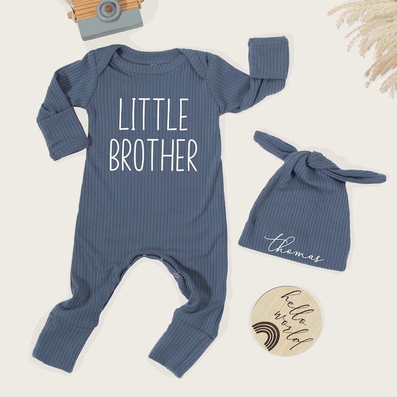 Pode incluir: Um conjunto de roupa de beb&eacute; azul canelado com as palavras "LITTLE BROTHER" estampadas na frente. O conjunto inclui um chap&eacute;u de n&oacute; a condizer com o nome "Thomas" e um disco de madeira que diz "hello world".