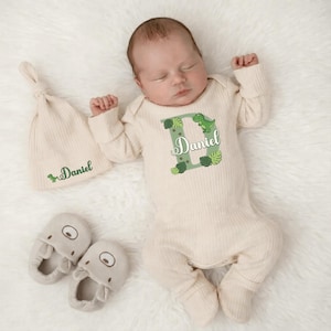 May include: A newborn baby wearing a cream-colored onesie, hat, and booties. The onesie features a green dinosaur and the name "Daniel" in a decorative font. The hat also has the name "Daniel" embroidered on it. The booties are light gray.