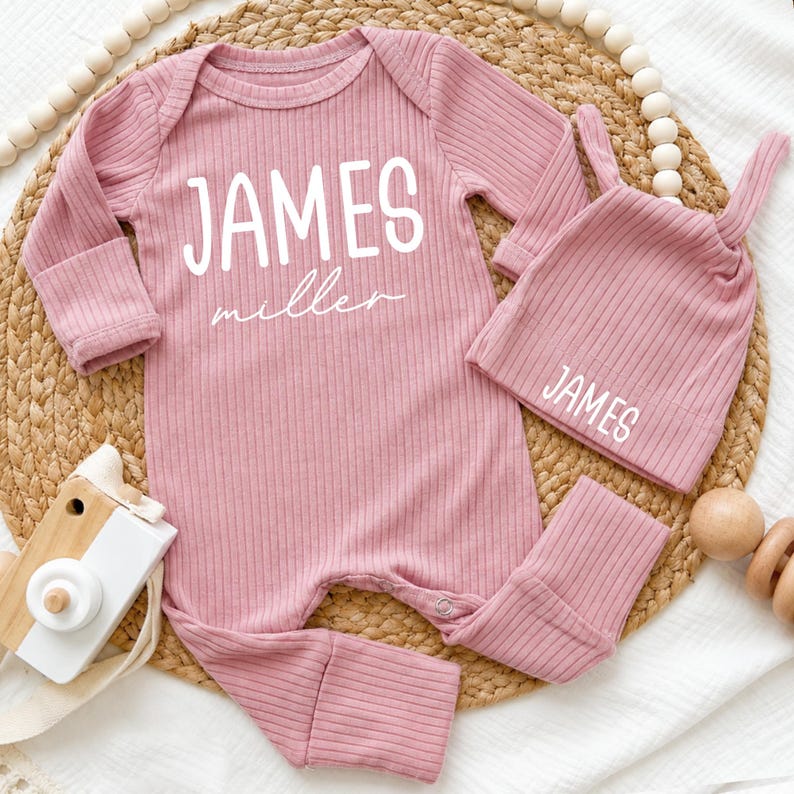 Pu&ograve; includere: Un set per neonati in tessuto a coste color rosa antico, composto da una tutina a maniche lunghe e un berretto con nodo. La tutina presenta la scritta bianca JAMES in stampatello sopra la parola miller in corsivo. Il berretto riporta la scritta JAMES.