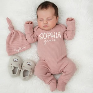 Pu&ograve; includere: Un neonato indossa un completo in maglia a coste rosa, cappello e pantofole. Il completo ha il nome "Sophia Grace" stampato sul davanti. Il cappello &egrave; ricamato con il nome "Sophia". Le pantofole sono grigio chiaro.