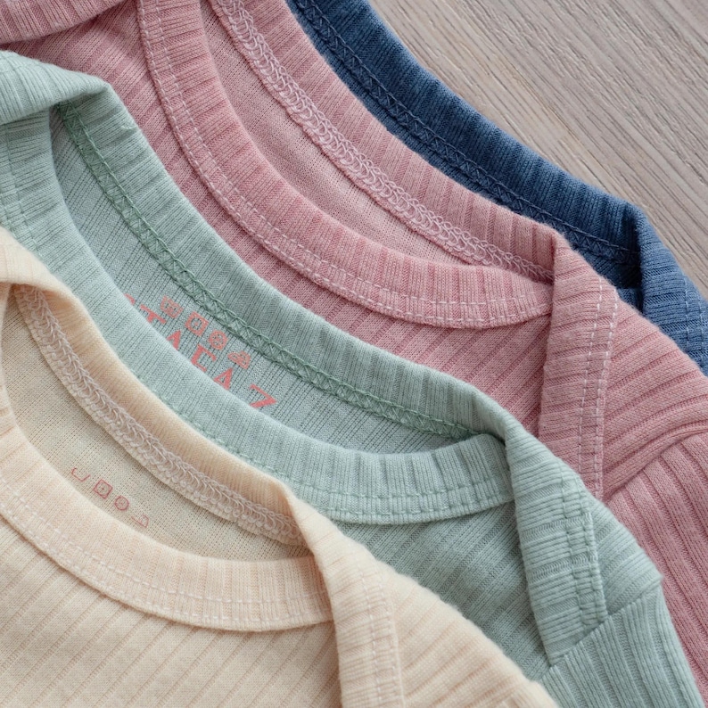 Pu&ograve; includere: Una pila di quattro body in cotone a coste nei colori crema, verde salvia, rosa antico e blu ardesia. Ogni capo presenta uno scollo rotondo con cuciture rinforzate e un'etichetta stampata visibile all'interno del colletto.