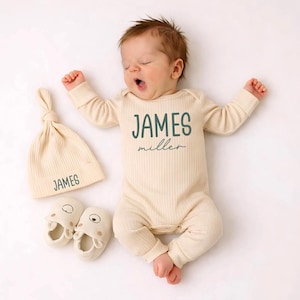 Pu&ograve; includere: Un body a maniche lunghe a coste color crema con il nome JAMES miller stampato in verde scuro, un berretto a nodo a coste abbinato con il nome JAMES e un paio di scarpine per neonati color crema con piccole orecchie da orso.