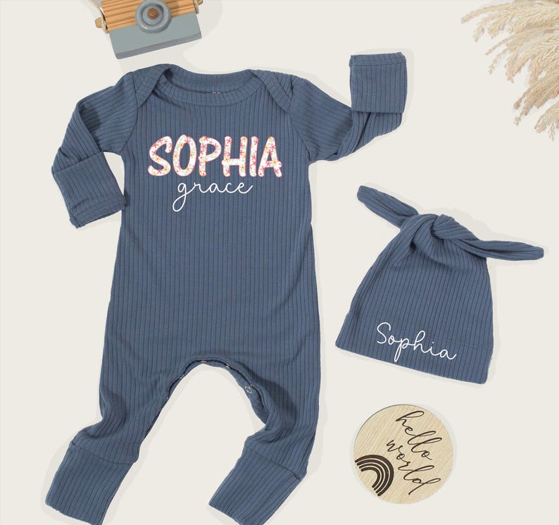 Pu&ograve; includere: Un completo per neonati in maglia a coste blu, composto da una tuta a maniche lunghe e un cappello annodato. La tuta presenta il nome "Sophia grace" in un design floreale e il cappello &egrave; ricamato con "Sophia". &Egrave; incluso anche un disco di legno con "hello world".