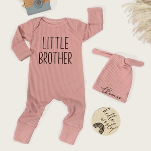 Pode incluir: Um conjunto de roupa de beb&eacute; rosa claro, incluindo um macac&atilde;o canelado com "LITTLE BROTHER" impresso na frente, um chap&eacute;u de n&oacute; a condizer com um nome e um disco de madeira com um arco-&iacute;ris e o texto "hello world". Uma c&acirc;mara de brinquedo est&aacute; no fundo.