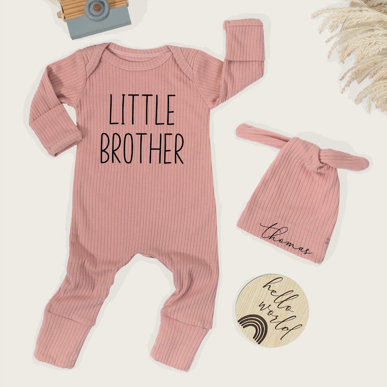Pode incluir: Um conjunto de roupa de beb&eacute; rosa claro, incluindo um macac&atilde;o canelado com "LITTLE BROTHER" impresso na frente, um chap&eacute;u de n&oacute; a condizer com um nome e um disco de madeira com um arco-&iacute;ris e o texto "hello world". Uma c&acirc;mara de brinquedo est&aacute; no fundo.