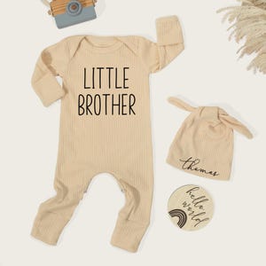 Pode incluir: Um conjunto de roupa de beb&eacute; em malha canelada bege. O conjunto inclui um body de manga comprida com "LITTLE BROTHER" estampado na frente, um chap&eacute;u com n&oacute; com um nome e um disco de madeira com "hello world" e um arco-&iacute;ris.