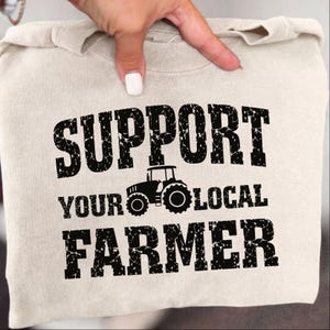 May include: A beige sweatshirt with the text "SUPPORT YOUR LOCAL FARMER" in distressed black lettering. A black tractor graphic is centered between "YOUR" and "LOCAL". The sweatshirt is folded and held by a hand.