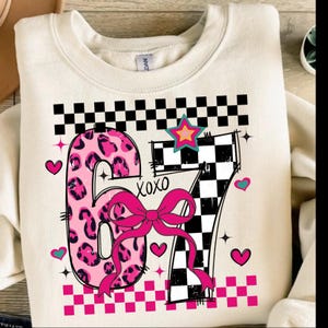 Könnte beinhalten: Cremefarbenes Sweatshirt mit einem grafischen Design mit der Zahl 67. Die Zahl 6 ist rosa mit Leopardenmuster, und die 7 ist schwarz-weiß kariert. Eine rosa Schleife und das Wort "xoxo" sind ebenfalls enthalten.