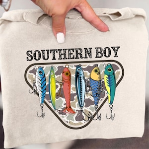 May include: A beige sweatshirt featuring the text SOUTHERN BOY in black letters above a triangular graphic. The graphic contains a camouflage background and six colorful fishing lures with hooks, rendered in blue, yellow, red, and green tones.