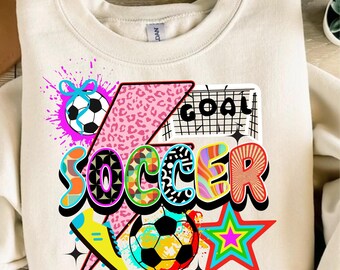 May include: Cream-colored sweatshirt with a colorful soccer-themed graphic. The design includes the word "SOCCER" in vibrant, multi-colored letters, soccer balls, a soccer cleat, and a star. The word "GOAL" is also featured.