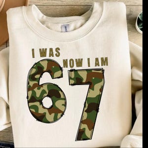 May include: Cream-colored sweatshirt with the text "I WAS NOW I AM" in brown. The number "67" is prominently displayed in a camouflage pattern, outlined in black. A casual, comfortable garment.