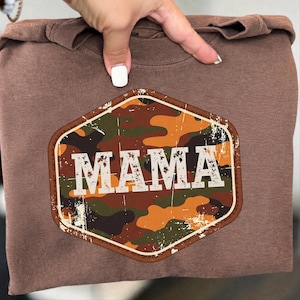 Może przedstawiać: Brązowa bluza z kapturem z wytartą naszywką w kamuflażu z napisem "MAMA" w białych literach. Naszywka ma kształt sześciokąta z brązową obwódką. Bluza jest w jednolitym brązowym kolorze.