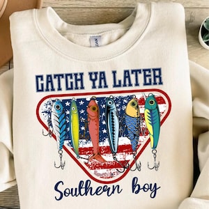 May include: Cream-colored sweatshirt with the text "CATCH YA LATER" above a graphic of six colorful fishing lures. The lures are set against a red, white, and blue background with the words "Southern boy" below.