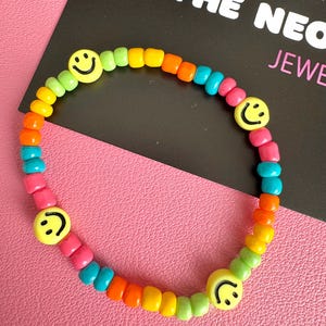 May include: A vibrant beaded bracelet in a rainbow gradient of colours, featuring yellow, green, orange, pink, and blue beads. The bracelet includes four yellow smiley face beads. The background includes the text "THE NEON JEWELRY".