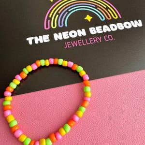May include: A vibrant beaded bracelet featuring neon pink, orange, yellow, and green beads. The bracelet is displayed on a pink surface, with a black card in the background. The card reads "THE NEON BEADBOW JEWELLERY CO." with a rainbow and stars graphic.