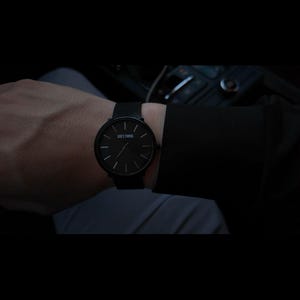 May include: A black wristwatch with a round face and minimalist design. The watch features a black band and a black dial with the text "GOD'S TIMING". The watch is worn on a wrist.