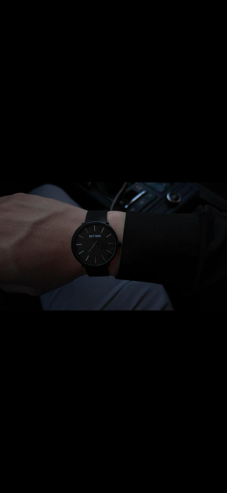May include: A black wristwatch with a round face and minimalist design. The watch features a black band and a black dial with the text "GOD'S TIMING". The watch is worn on a wrist.