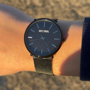 May include: A black wristwatch with a dark brown face and a mesh band. The watch face has the text "GOD'S TIMING." The watch has silver-colored hour markers and hands. The watch is on a wrist.