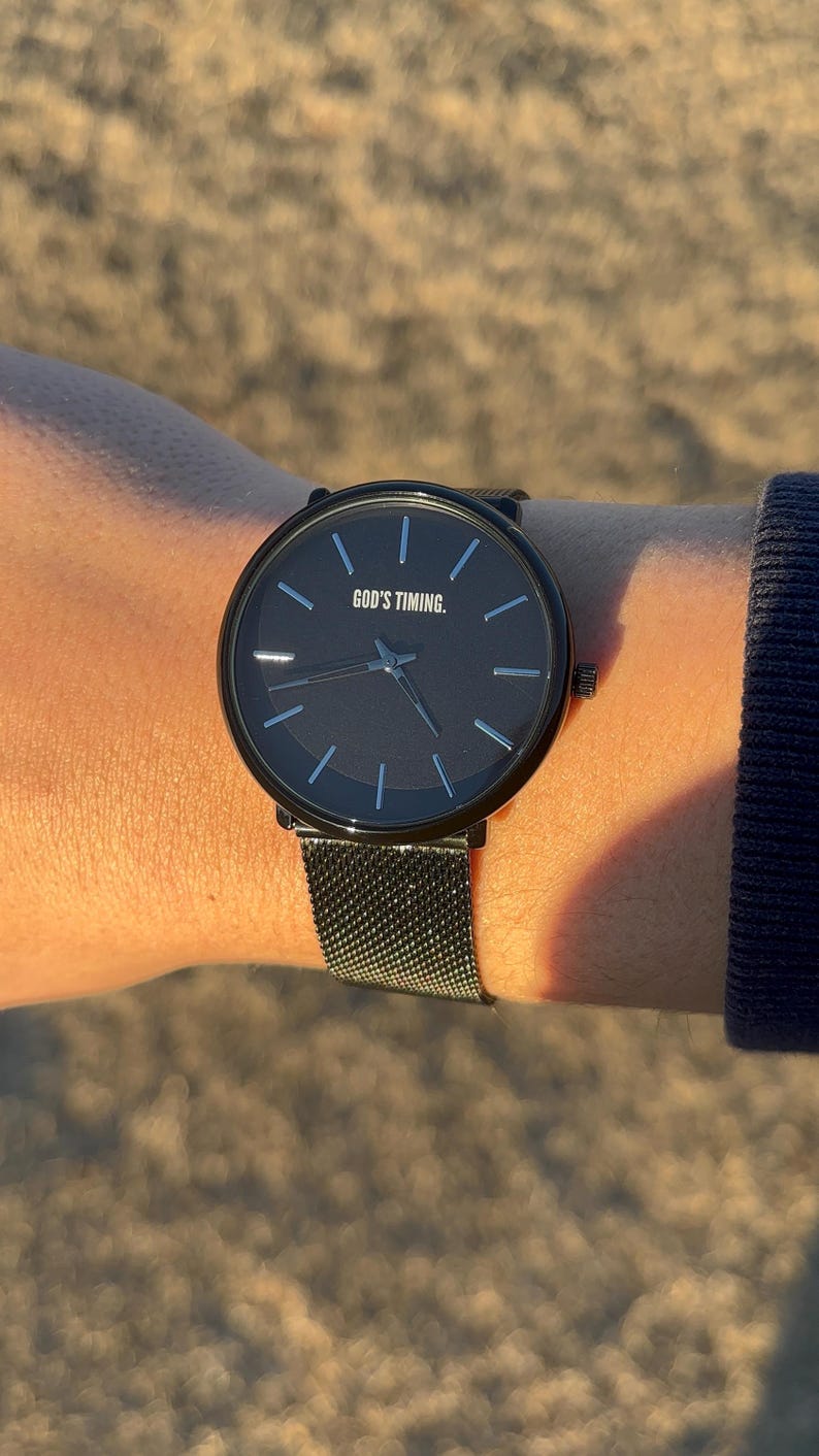 May include: A black wristwatch with a dark brown face and a mesh band. The watch face has the text "GOD'S TIMING." The watch has silver-colored hour markers and hands. The watch is on a wrist.