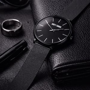 May include: A black wristwatch with a mesh band, featuring a black dial with white markers and the text "GOD'S TIMING." The watch is displayed with a black wallet, pen, ring, and sunglasses, all on a textured surface.