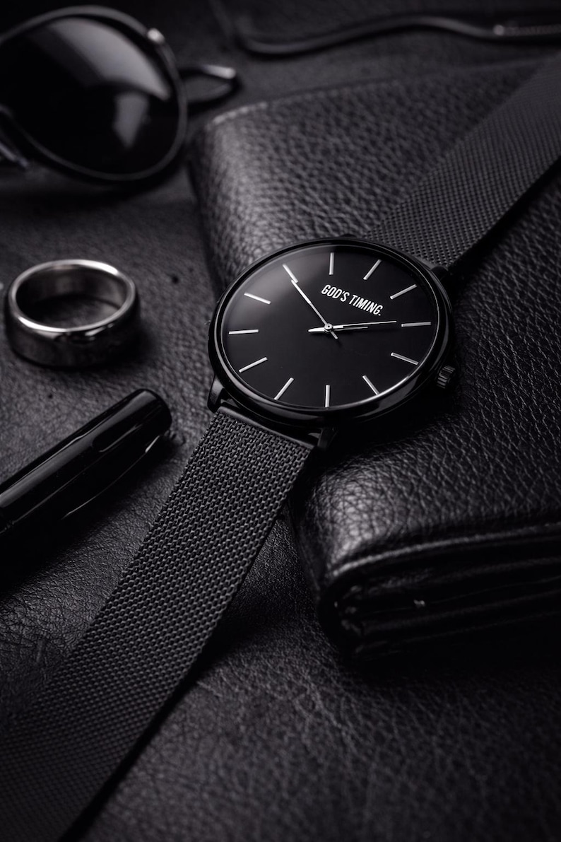 May include: A black wristwatch with a mesh band, featuring a black dial with white markers and the text "GOD'S TIMING." The watch is displayed with a black wallet, pen, ring, and sunglasses, all on a textured surface.