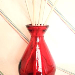 May include: A red glass vase with a bulbous base and a narrow neck, filled with light-colored reed diffuser sticks. The vase sits against a backdrop of a white cloth with colorful stripes.