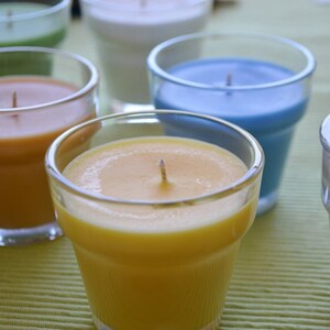 May include: Five glass candle jars with different colored waxes. The jars are arranged in a row on a green and white striped surface. The waxes are yellow, orange, green, blue, and brown.