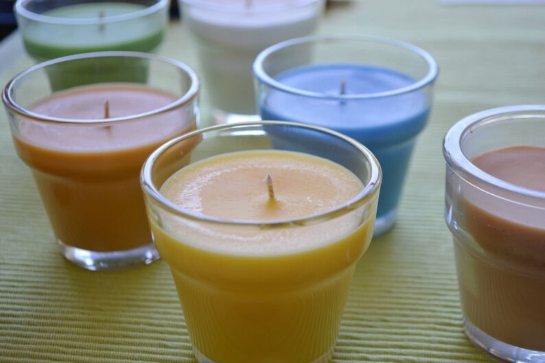 May include: Five glass candle jars with different colored waxes. The jars are arranged in a row on a green and white striped surface. The waxes are yellow, orange, green, blue, and brown.