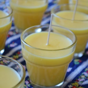 May include: A close-up of several yellow candles in glass jars with wicks. The candles are arranged on a blue and white patterned fabric.