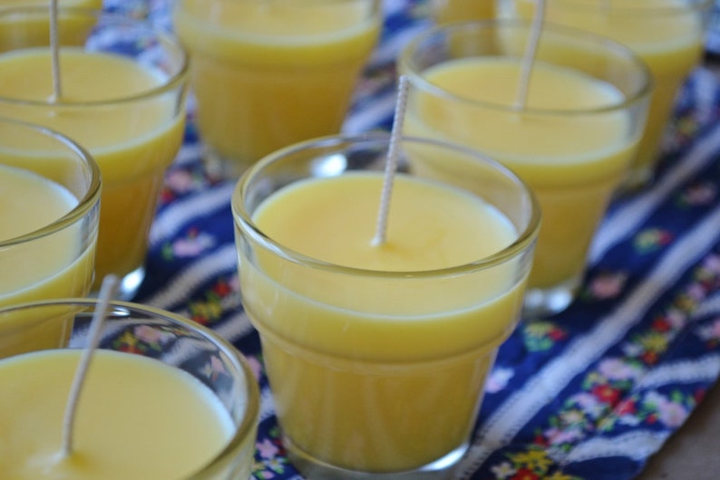May include: A close-up of several yellow candles in glass jars with wicks. The candles are arranged on a blue and white patterned fabric.