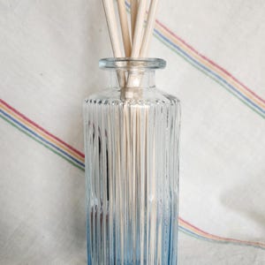 May include: A clear glass diffuser bottle with a blue ombre effect at the base, filled with wooden reeds. The bottle has vertical ribbed detailing. The background is a white cloth with colorful stripes.