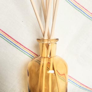 May include: Amber glass diffuser bottle filled with wooden reeds. The bottle has a rounded shape and a narrow neck. The background is a white fabric with colorful stripes. The reeds are light brown and extend upwards from the bottle.