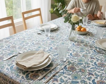 May include: A dining table set for a meal, featuring a floral patterned tablecloth in blue, green, and peach hues. The table is set with white plates, silverware, and clear drinking glasses. A vase of flowers and a bowl of oranges are also present.