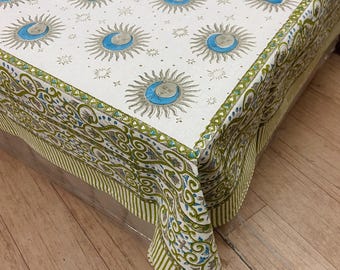 May include: Cream-colored tablecloth with a celestial pattern featuring suns, moons, and stars in shades of gray and blue. The border has a green and white striped design. The tablecloth is made of a soft, durable fabric.
