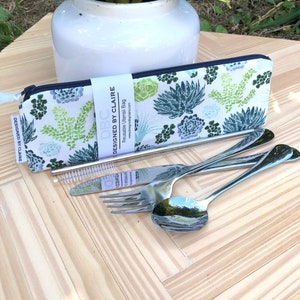 May include: A white zippered pouch with a green and blue floral pattern. The pouch is labeled "Designed by Claire" and contains a set of stainless steel utensils, including a fork, knife, spoon, and straw with a cleaning brush.