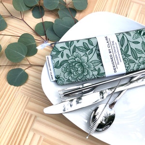 May include: A green floral reusable utensil bag with the text "Designed by Claire" and "Reusable Utensil Bag" on the front. The bag is open and a set of silver utensils are on a white plate.