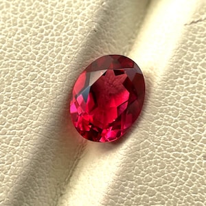 May include: A faceted, oval-shaped, deep red gemstone. The gem has a brilliant cut, reflecting light and showcasing its rich colour. The stone is set against a textured, off-white surface.