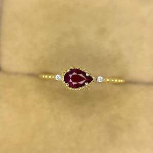 Può includere: Un anello in oro con un rubino rosso centrale a forma di pera circondato da un bordo metallico perlato. Due piccole pietre d'accento rotonde e trasparenti sono incastonate sulla fascia, una su ciascun lato del rubino. La fascia ha una finitura.