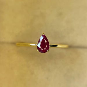 Pode incluir: Um anel de ouro com uma gema de rubi vermelho escuro em forma de pêra. O rubi é o ponto focal, cravado em uma delicada banda de ouro. O anel é exibido em um fundo neutro, destacando a cor e a clareza da gema.